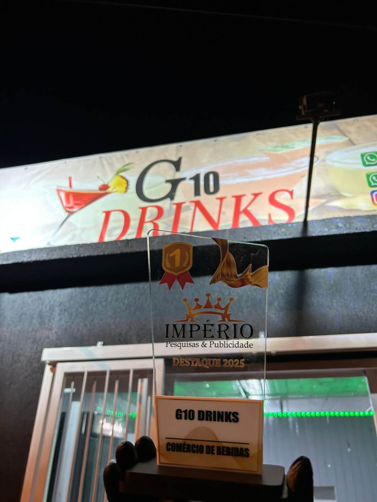 Troféu Império Pesquisas e Publicidade em frente à placa do G10 Drinks